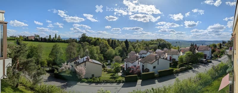 A scenic view of a residential area with houses, trees, and a clear blue sky, featuring a distant landscape and rolling hills.