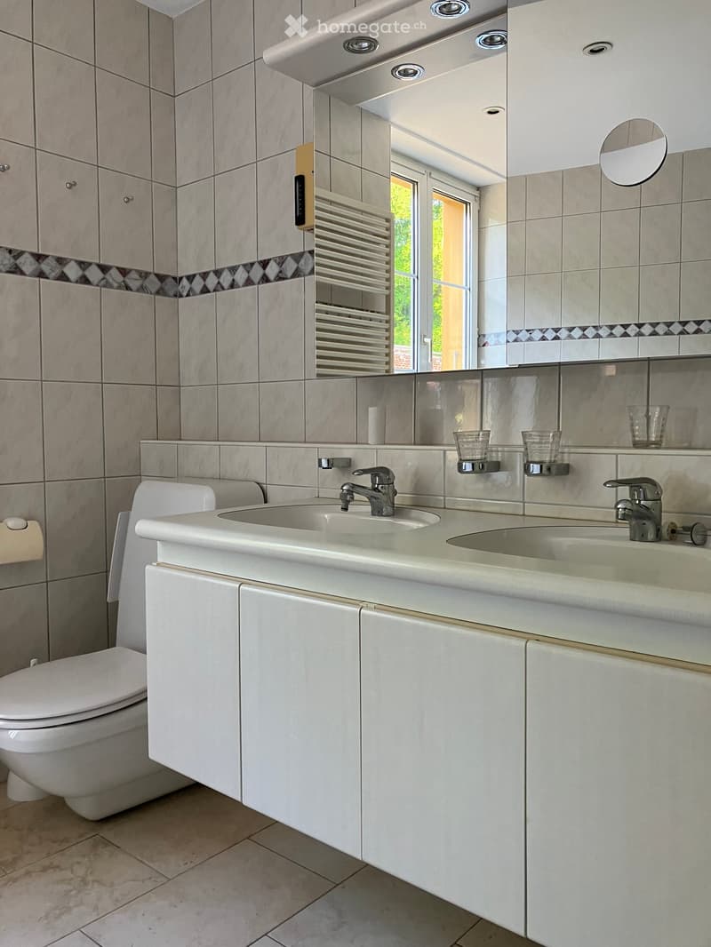 Modern bathroom with double sink, toilet, mirror, and radiator. Light gray tiles and white cabinetry.