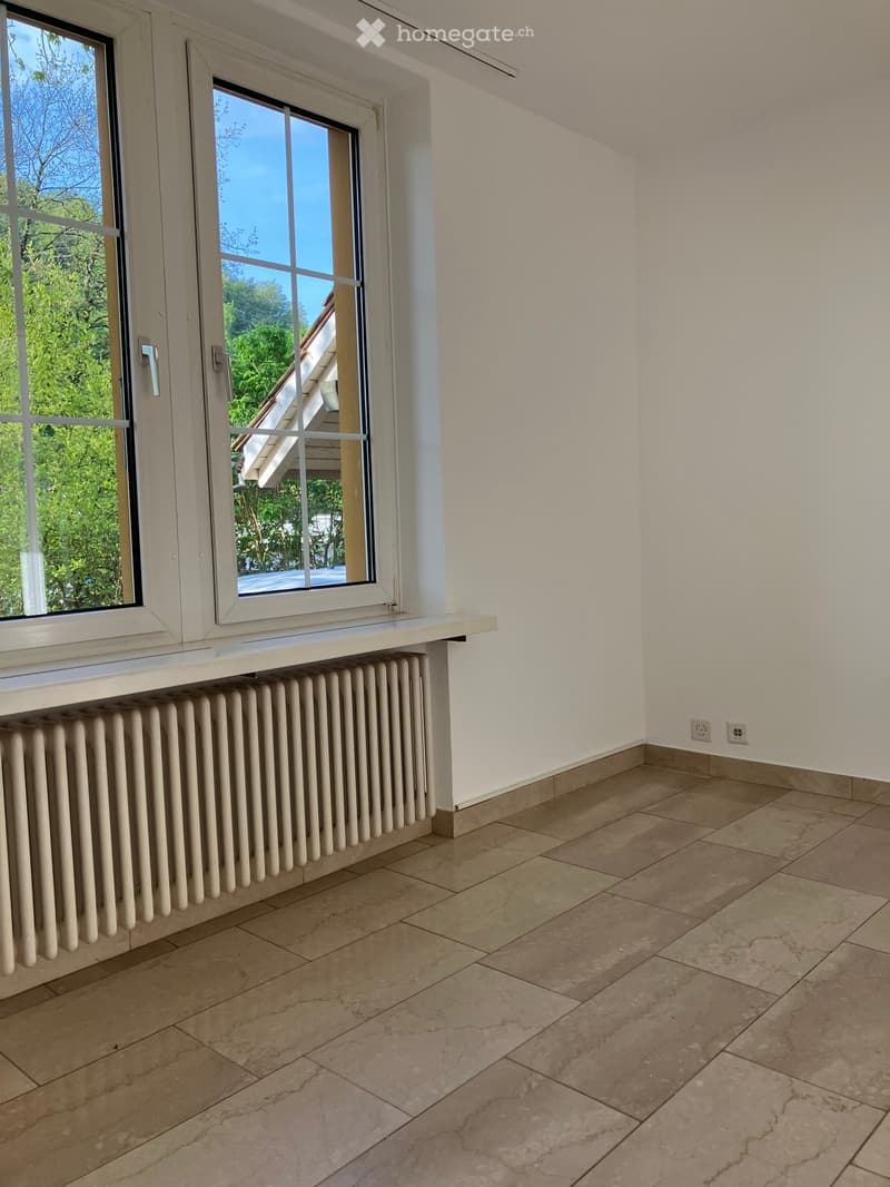 Empty room with tiled floor, white walls, radiator, and large windows with a view of greenery outside.