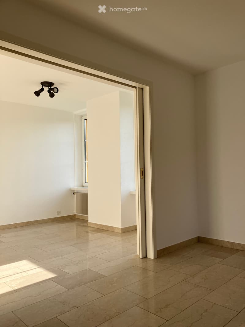 Empty room with beige tiled flooring, white walls, and a large open doorway leading to another space. A small window and radiator are visible in the adjacent area.