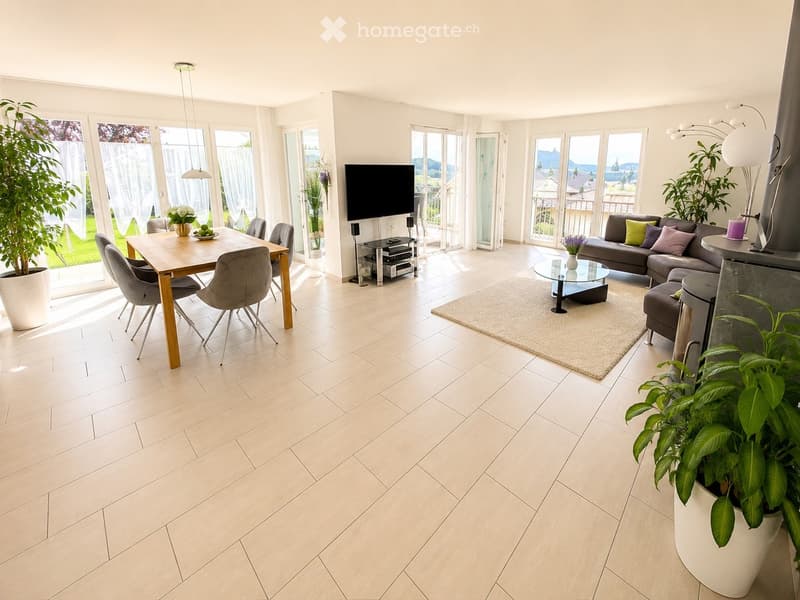 Bright, open living and dining area with large windows and sliding doors leading to a balcony. Features a grey sectional sofa, glass coffee table, wooden dining table with four chairs, and a television on a stand. The space includes modern lighting, potte