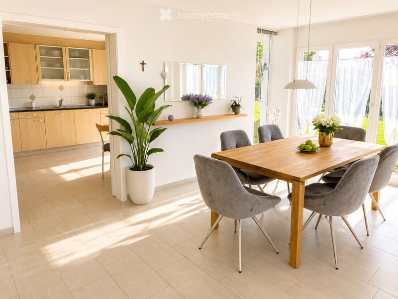 Bright dining area with a wooden dining table surrounded by grey upholstered chairs, a large potted plant, and a modern kitchen in the background.