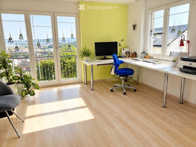 Bright office space with wooden flooring, large windows offering a scenic view, a desk with a computer screen, a blue office chair, and a printer, complemented by potted plants and a comfortable grey armchair.