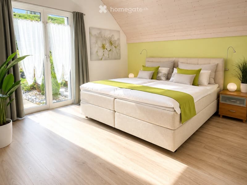 Bright bedroom with a large bed, green and white bedding, nightstand with lamp, potted plant, and a view of a garden through large windows.
