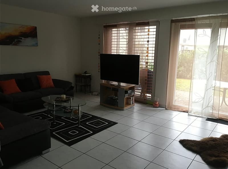 Living room with tiled floor, dark grey sectional sofa with orange cushions, glass coffee table, TV on stand, small side table, and area rug.