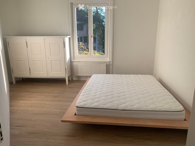 A furnished bedroom with a wooden floor, a white wardrobe, a bed with a mattress, and a window with a radiator below it.