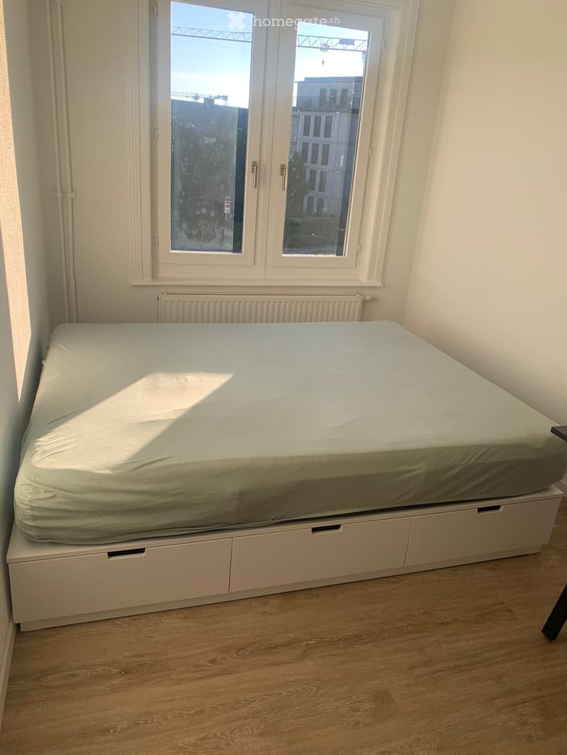 A simple bedroom with a single bed, light-colored bedding, and a radiator beneath a window. The floor is wooden, and the walls are white.