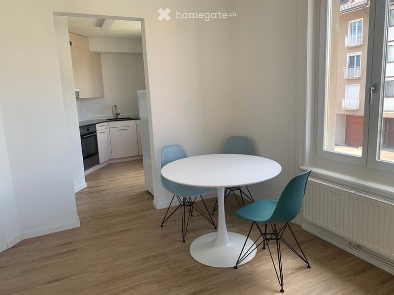 A modern, furnished room with a round white table and four blue chairs, a small kitchen area with an oven, sink, and refrigerator, and large windows offering a view of the street.