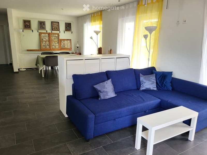 Modern living room with a blue sectional sofa, white coffee table, dining area with chairs and table, and decorative wall shelves.