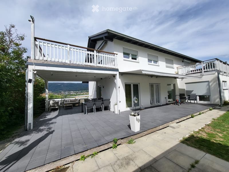 Modern two-story house with white exterior, multiple balconies, and a spacious wooden deck area furnished with seating and a dining set.