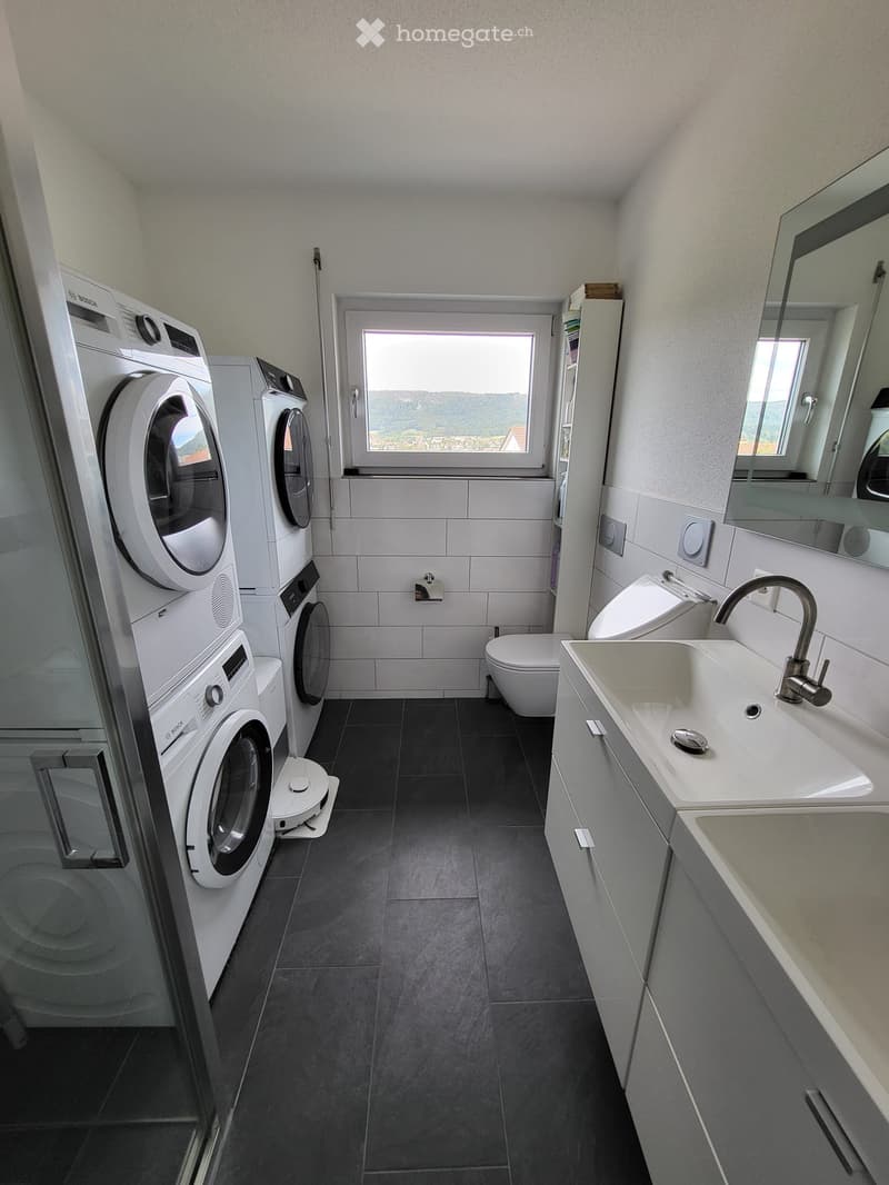 Modern bathroom with dark tiled floor, white walls, and large window. Includes a washing machine, dryer, sink with mirror, and toilet.