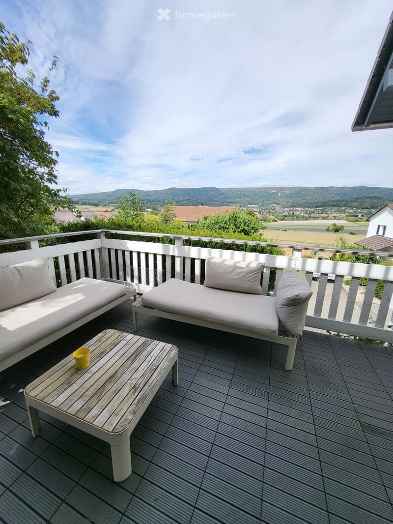Balcony with wooden decking, white railing, two white sofas, small wooden table with a yellow cup, scenic countryside view.