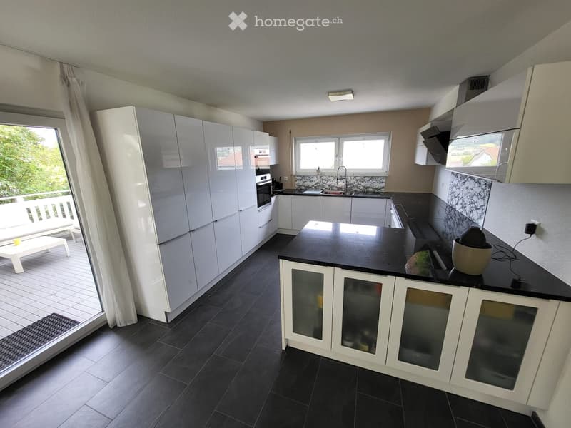 Modern kitchen with white cabinets, black countertops, and a large window overlooking a balcony. Features include a sink, faucet, oven, and a small white stool near the window.