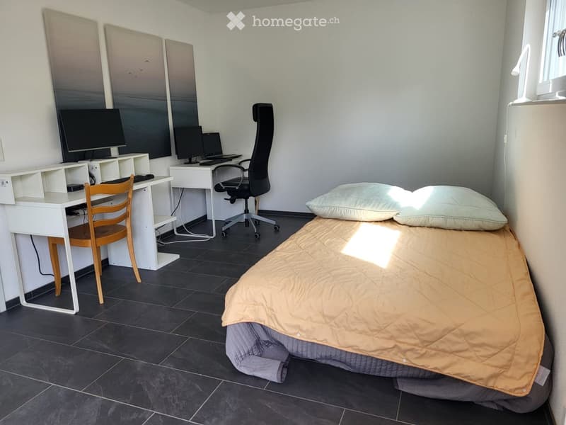 A furnished room with a bed, desk, and chairs. The room features a tiled floor and a window letting in natural light.