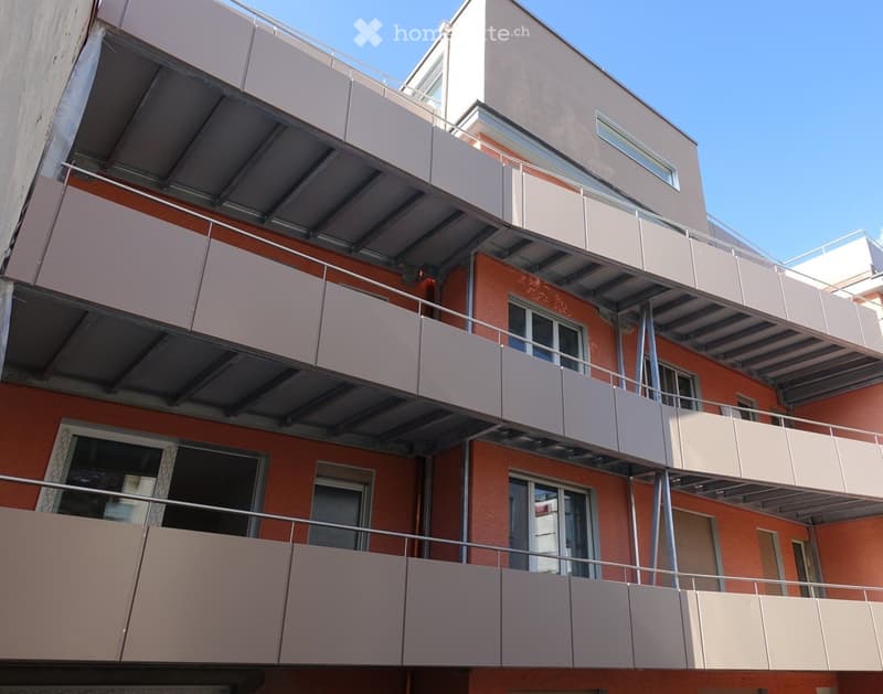 Modern multi-story building with orange brick facade, multiple balconies with white railings, and large windows.
