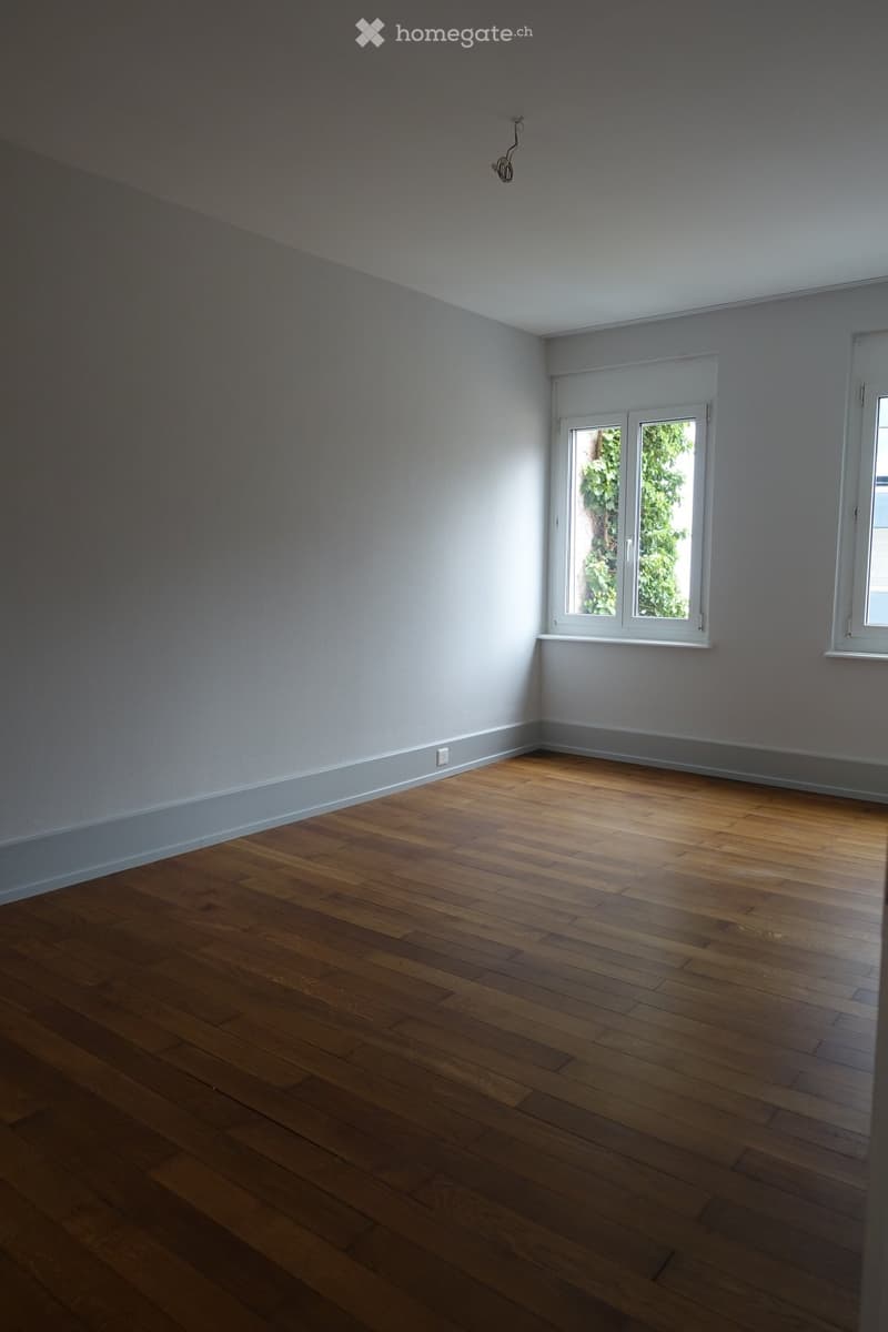 Empty room with wooden flooring and light grey walls, featuring two windows and a visible electrical outlet.