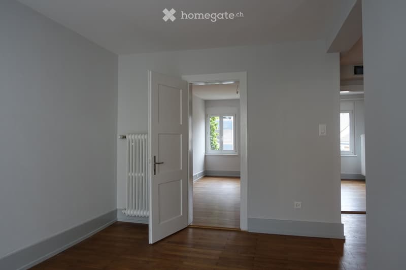 Empty hallway with light grey walls, wooden flooring, and a white radiator on the left side. An open door leads to another room with a window and wooden floor visible in the background.