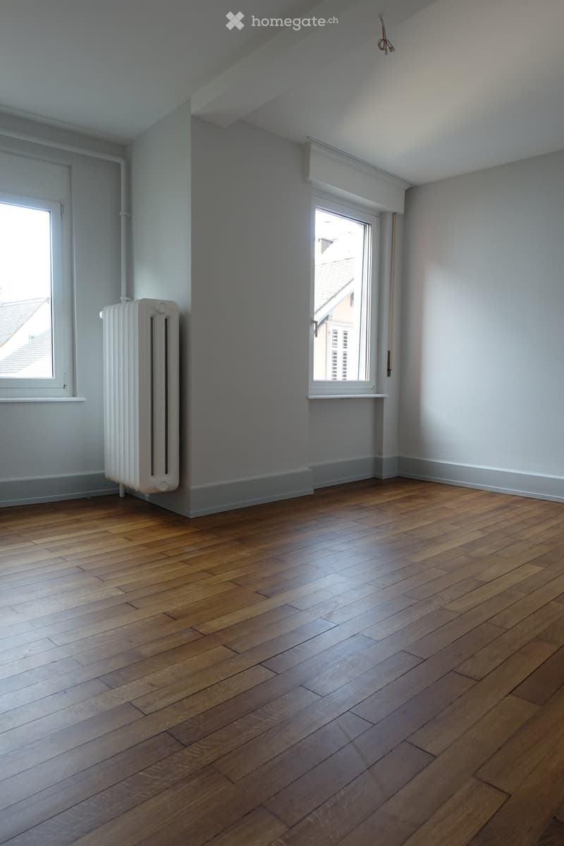 Empty room with wooden flooring, light grey walls, and two windows. A white radiator is mounted on one wall.