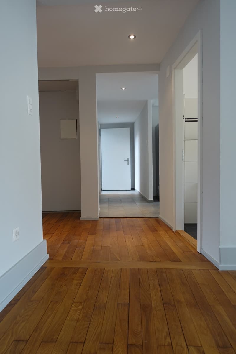 Empty hallway with wooden flooring, white walls, and recessed lighting. Features a closed door at the end and a partially visible room on the right.