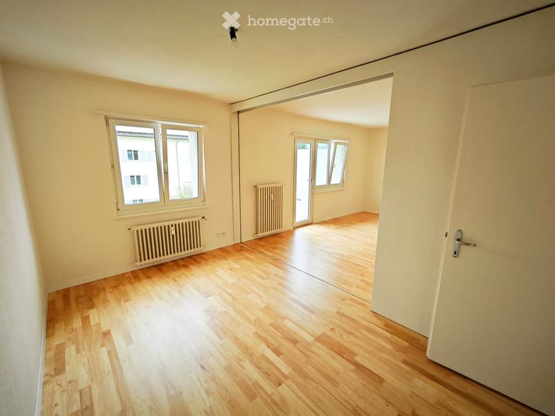 Empty room with wooden flooring, white walls, and two windows with radiators underneath. The room is connected to another space through an open doorway.