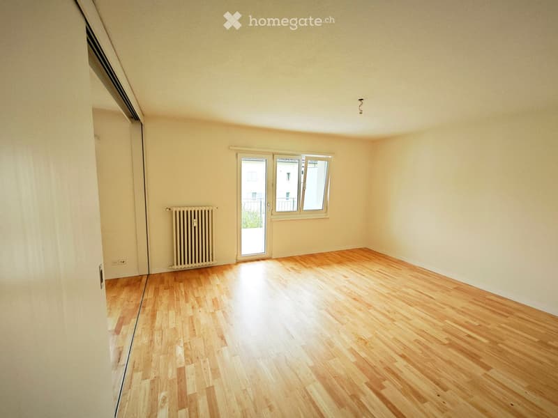 Empty room with wooden flooring, white walls, a radiator, and a large window with a sliding door leading to a balcony.