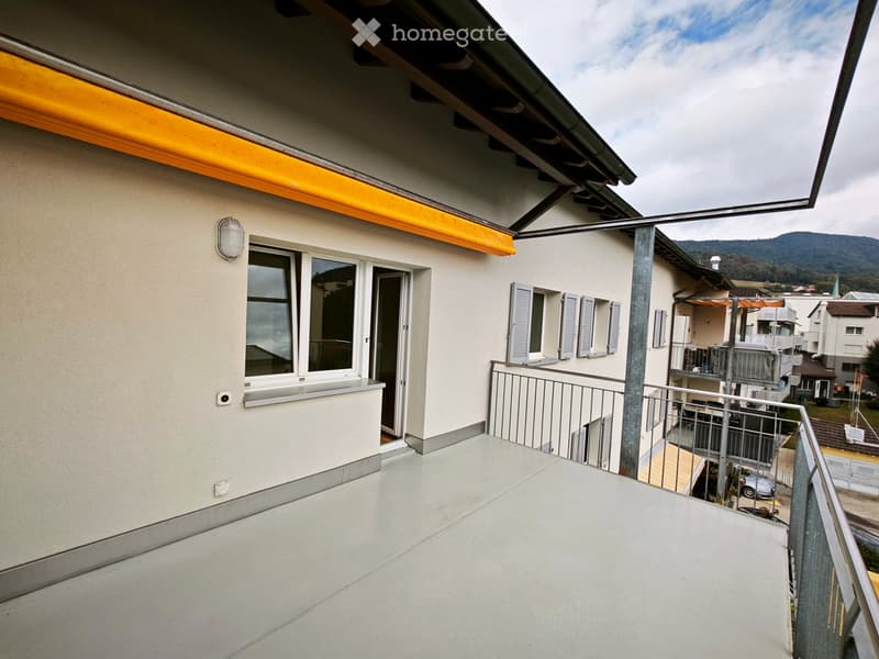 Balcony with a metal railing, a window, and a door leading to the interior. The view includes neighboring buildings and a mountainous landscape.