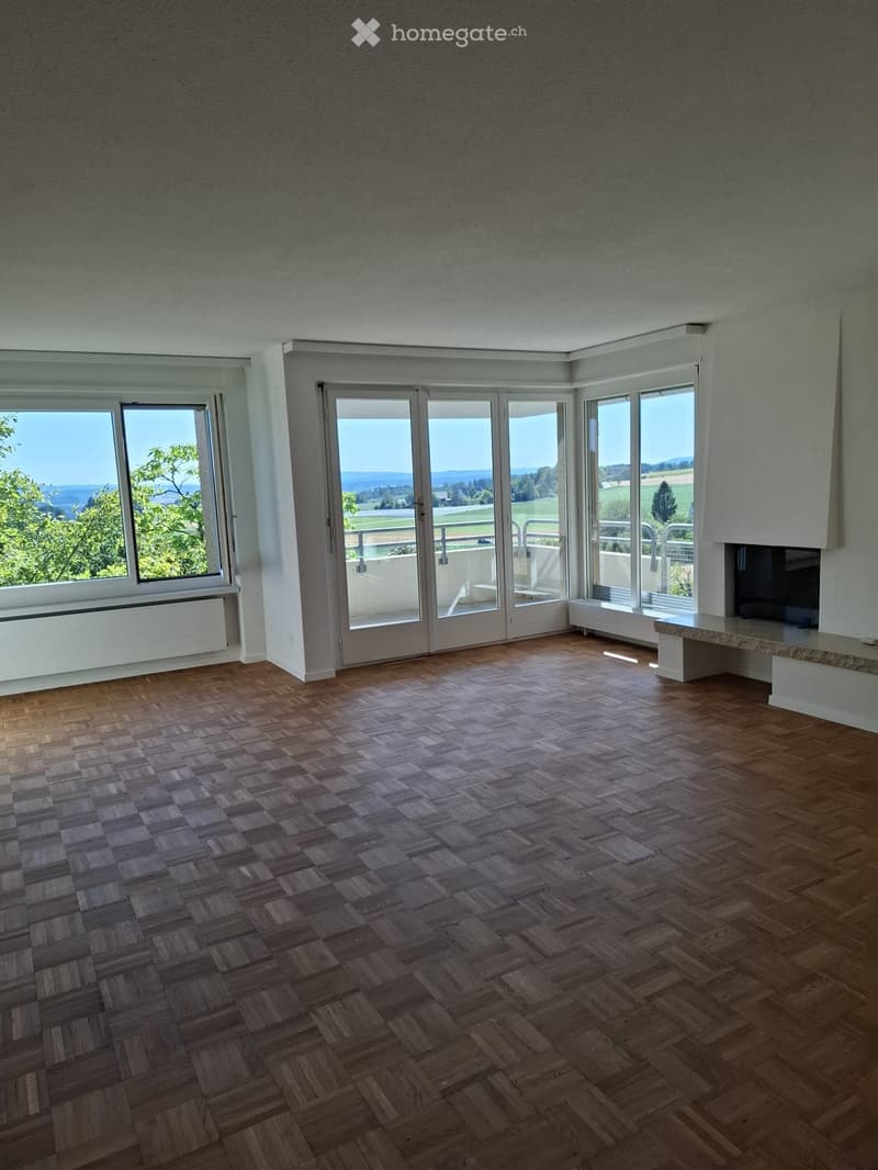 Empty room with wooden herringbone flooring, large windows and sliding glass doors leading to a balcony, offering scenic views of greenery and fields.