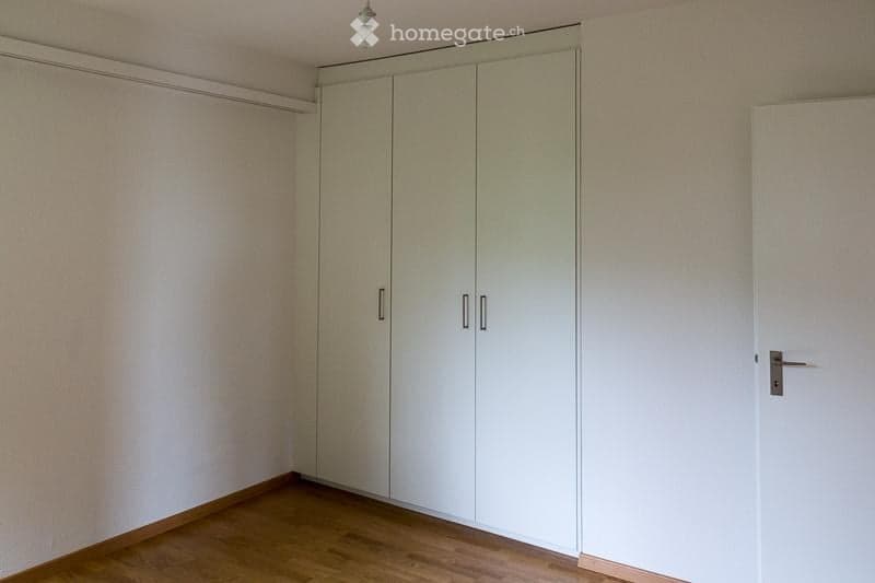 Empty room with light-colored walls, wooden flooring, and built-in white wardrobes.