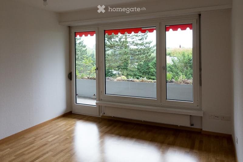 Empty room with wooden flooring, large windows with red awnings, and access to a balcony.