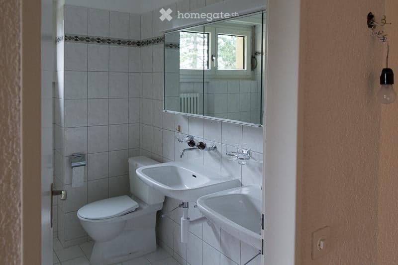 A small bathroom with white tiled walls and floor, featuring a toilet, two sinks, a large mirror above the sinks, and a window with a view of greenery outside.