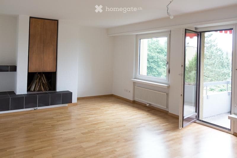 Empty living room with wooden flooring, white walls, large windows, and a sliding glass door leading to a balcony.