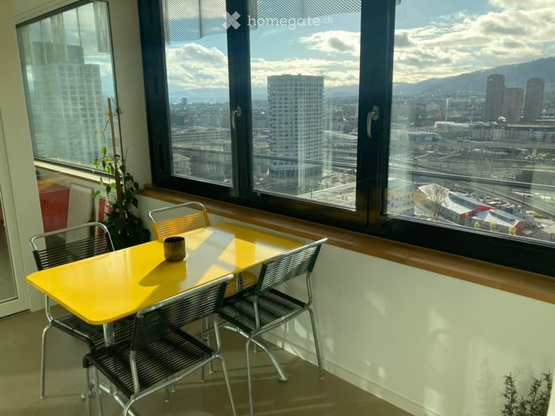 Bright room with large windows offering a city view, featuring a yellow table, chairs, and a cup.
