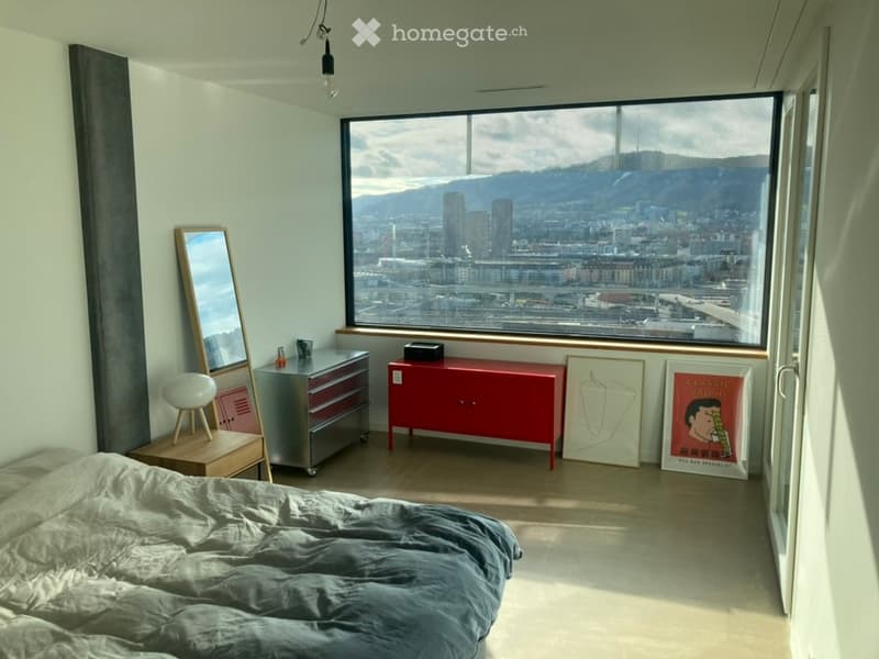 Bright bedroom with large window offering city and mountain view, featuring a bed, mirror, stool, cabinet, drawer unit, and framed artwork.