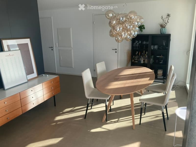 Brightly lit dining area with a round wooden table surrounded by chairs, a modern cabinet with picture frames, and a decorative chandelier hanging from the ceiling.