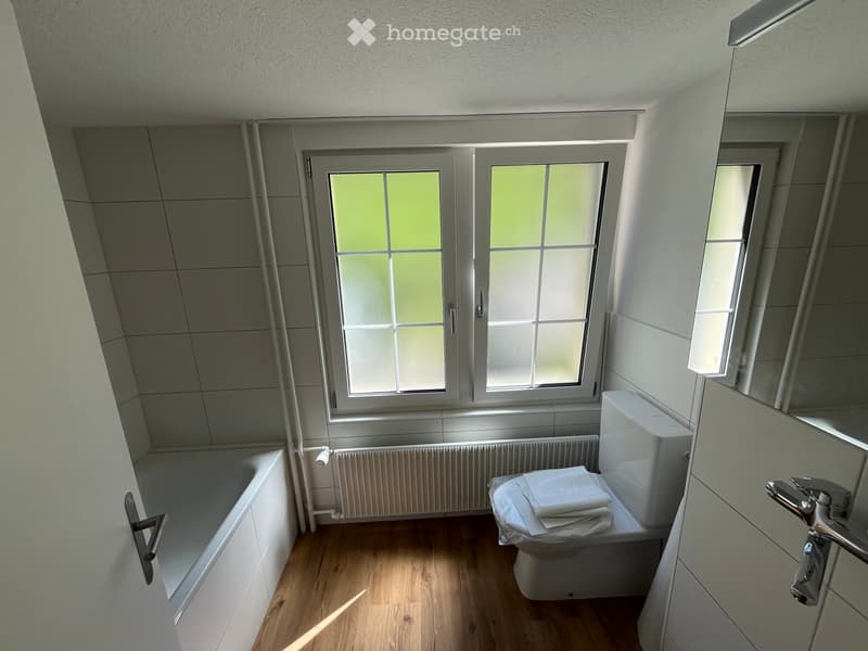 Modern bathroom with a bathtub, toilet, and towel on the toilet seat. The room features wooden flooring, white tiled walls, and large windows providing natural light.