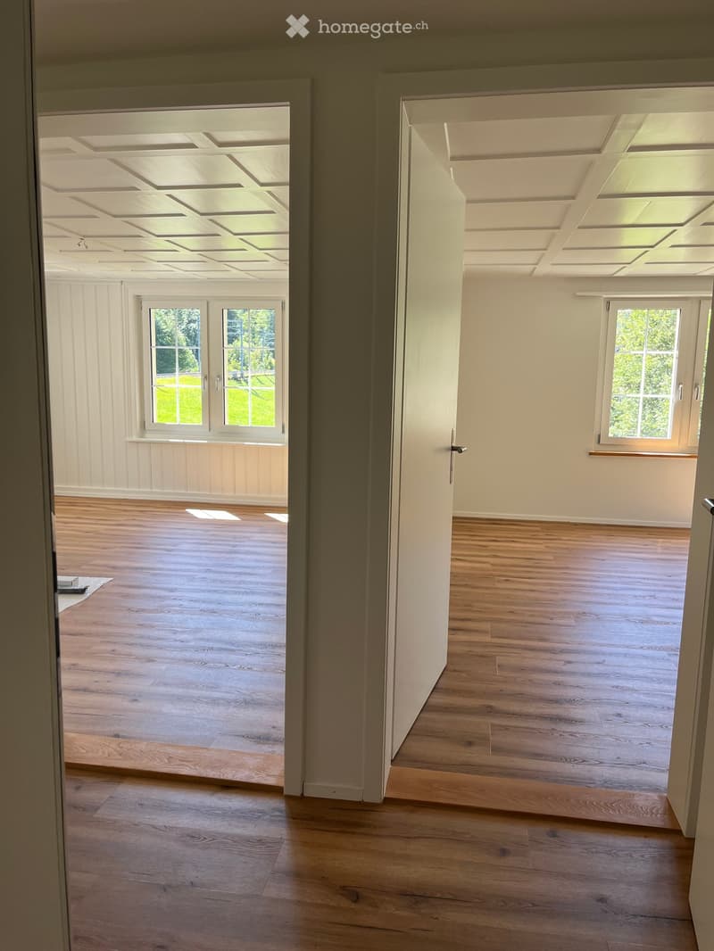 Empty room with wooden flooring, white walls, and large windows. The room has a high, coffered ceiling and is connected to another similar room through an open doorway.