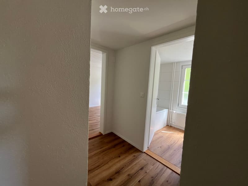 Empty hallway with wooden flooring, leading to a small room with a window and a radiator.