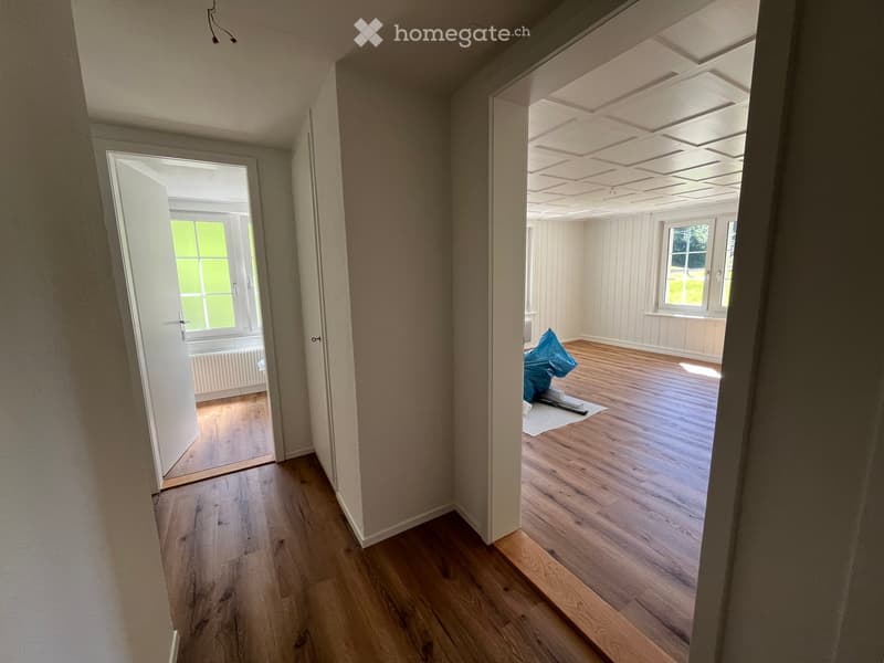 Empty hallway with wooden flooring, white walls, and an open doorway leading to another room with wooden flooring and white walls.