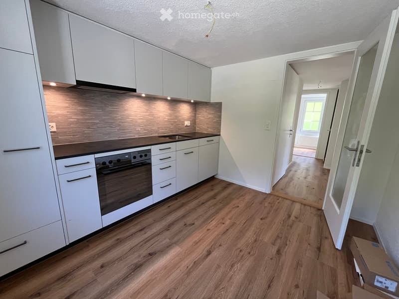 Modern kitchen with white cabinets, wooden flooring, and built-in oven. Open doorway leads to another room.