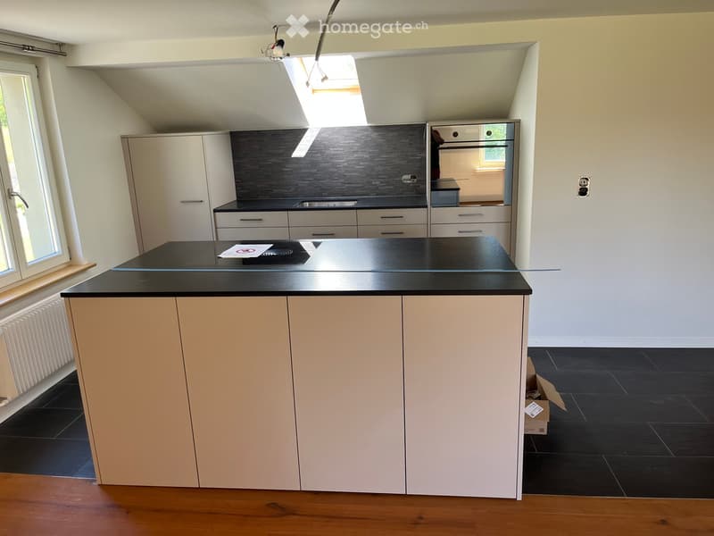 Modern kitchen with a central island, white cabinets, black countertops, and a built-in oven. The space features large windows and a skylight, with dark tiled flooring.