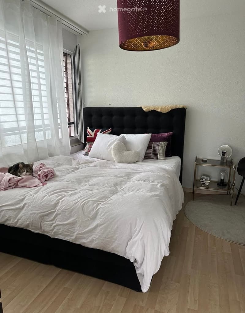 A modern bedroom with a large bed, headboard, multiple pillows, a nightstand with a mirror, and a chair. The room features wooden flooring and large windows with white blinds.