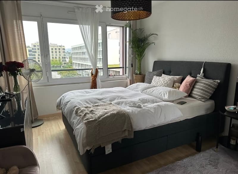 Modern bedroom with large windows offering a city view, a neatly made bed with pillows and blankets, a nightstand, a fan, a vase with flowers, and a small table with a chair.