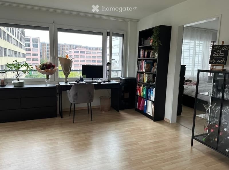 Bright, modern room with large windows, wooden floor, desk with computer, bookshelf, wine rack, and decorative items.