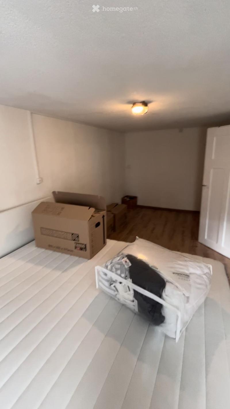 A partially furnished bedroom with a bed, pillow, and cardboard boxes stacked against the wall. The room has wooden flooring and white walls.