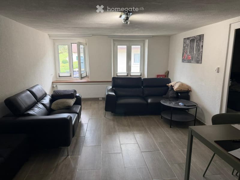Modern living room with two black leather sofas, a round coffee table, a small table, and a chair. The room features large windows and a painting on the wall.