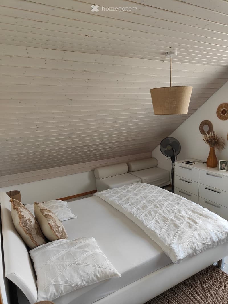 Cozy bedroom with a white bed, beige pillows, and a textured blanket. Features a nightstand with a fan, a vase with dried flowers, and a mirror on the wall. The room has wooden paneling and a hanging lamp.