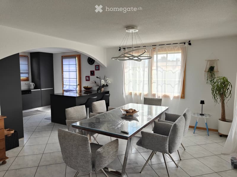 Modern dining area with a glass dining table surrounded by six grey upholstered chairs. The room features a sleek kitchen counter with cabinets, a vase with white flowers, and a decorative clock on the wall. Large windows with sheer curtains allow natural