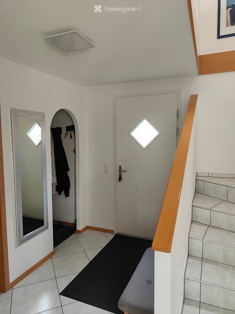 A clean, well-lit hallway with tiled flooring, a wooden handrail, a black runner, and a mirrored wall. A door leads to another room, and a coat rack is visible in the adjacent space.