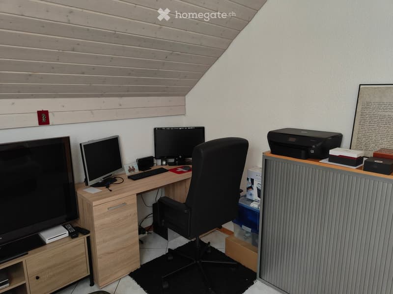 A modern office space with a wooden desk, ergonomic chair, multiple monitors, keyboard, mouse, printer, and a radiator. The room features a slatted wooden ceiling and a black rug under the chair.