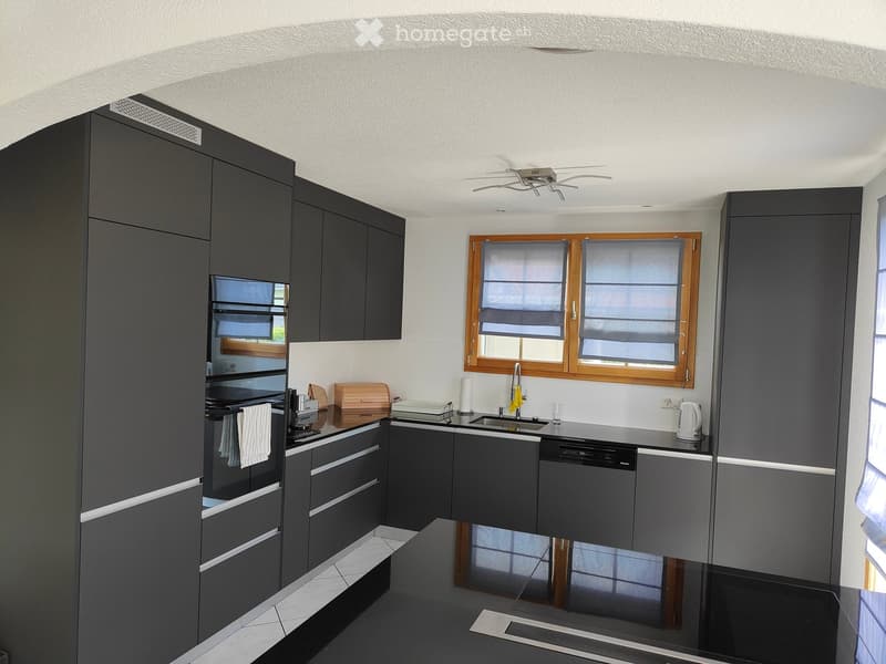 Modern kitchen with dark grey cabinets, stainless steel appliances, and a central island. Features include an oven, dishwasher, sink, and various small appliances.
